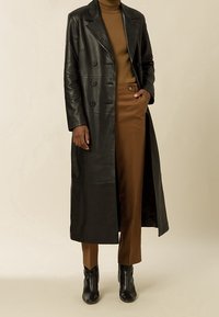 Černý kožený dvojřadý kabát s velkými knoflíky, spárovaný s hnědým rolákem a upravenými hnědými kalhotami. Černé kotníkové boty. Hladká textura.