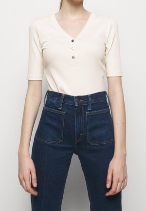 Kvinde iført en ribbet beige henley-top med korte ærmer og tre knapper, stoppet ind i højtaljede mørkeblå jeans med forlommer.