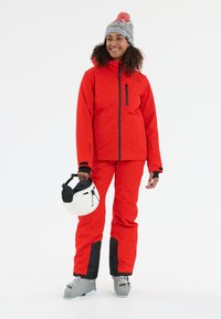 Rode waterdichte ski-jas en broek met een aangesloten model; grijze gebreide muts met pompon; witte skihelm; grijze skischoenen met accenten.