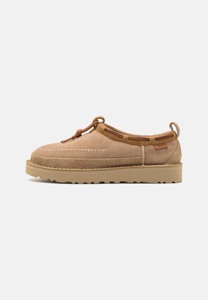 Chaussure à enfiler en daim marron clair avec un bout rond, présentant des coutures décoratives, des accents en cuir et une semelle en caoutchouc.
