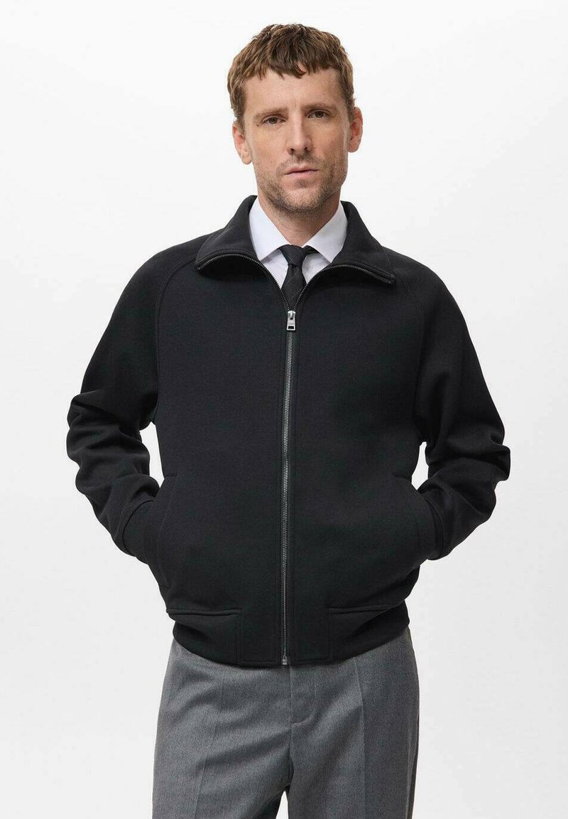 Chaqueta negra con cremallera y cuello alto, hecha de una tela suave, con dos bolsillos frontales y puños acanalados. Combinada con pantalones grises.