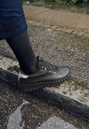 Piede con sneaker nero impermeabile che calpesta il bordo bagnato del marciapiede con una pozzanghera d'acqua vicina, all'aperto in una giornata piovosa.