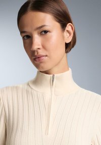 Krém színű, bordázott zipzáros pulóver magas gallérral, keskeny cipzárral és texturált anyaggal. Közeli nézet a nyakrészről és az arcról.