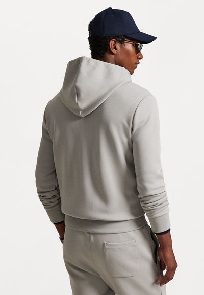 Sweatshirt cinza com capuz, punhos e bainha canelados. Textura suave e macia. Usado com calças a condizer e boné azul-marinho, mostrando a vista traseira.