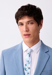 Completo azzurro chiaro con finitura strutturata, camicia bianca e cravatta floreale con tonalità di blu. Revers ordinato e capelli curati.