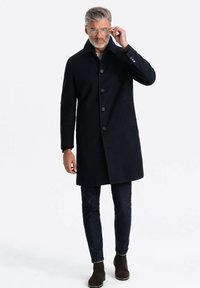 Cappotto di lana blu scuro con colletto alto, caratterizzato da cinque bottoni, una tasca laterale e una vestibilità sartoriale, abbinato a jeans scuri e stivali marroni.
