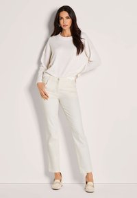 Vrouw met lang donker haar, gekleed in een witte trui, crèmekleurige rechte broek en beige hakken met loafers, staande tegen een lichte muur.