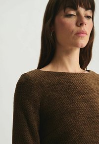 Pull en laine marron texturé avec un décolleté carré ; il présente un motif tricoté et des manches longues. Design minimaliste avec une finition lisse.
