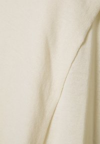 Světle béžová pletená látka s hladkou texturou, zdobená zavěšeným designem a jemným žebrováním, které zdůrazňuje měkkost a flexibilitu.