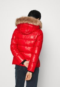 Rød puffjakke med en blank finish, der har en pelsforet hætte, quiltet design og ribbede manchetter. Bagsiden har vandrette sømme.