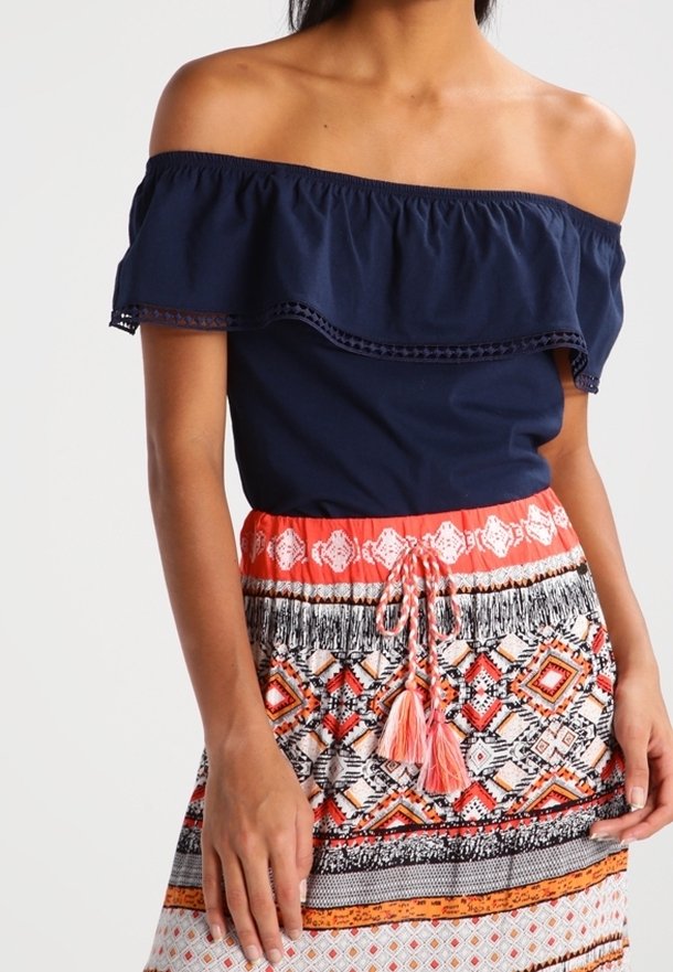 Mörkblå off-the-shoulder topp med volangkrage, kombinerad med en mönstrad kjol med korallfärgade, svarta och vita geometriska mönster samt tofsdetaljer.