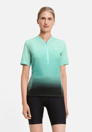 Woman standing wearing a short-sleeve turquoise gradient cycling jersey with a front zipper and black padded cycling shorts.