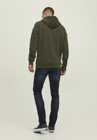Grüne Kapuzenjacke mit Kängurutasche, gerippten Bündchen und Saum, kombiniert mit dunklen Jeans und navyfarbenen Sneakers vor einem neutralen Hintergrund.