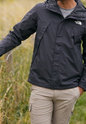 Person trägt eine schwarze North Face Jacke und beige Hose und läuft durch ein grasbewachsenes Feld, wobei sie mit einer Hand hohes Gras berührt.