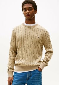 Beige kabelsweater met geribde boorden en taille, met een klein logo op de borst, gedragen over een wit T-shirt.