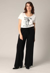 Zwarte wijde broek met een gestructureerde afwerking, gecombineerd met een witte T-shirt met zwarte grafische tekst. Model draagt zwarte enkellaarsjes.