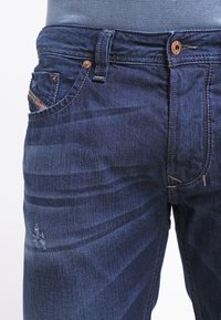 Jeans in denim blu scuro con design aderente, leggere sfumature sbiadite, bottoni di rame e un piccolo dettaglio usurato sulla gamba.