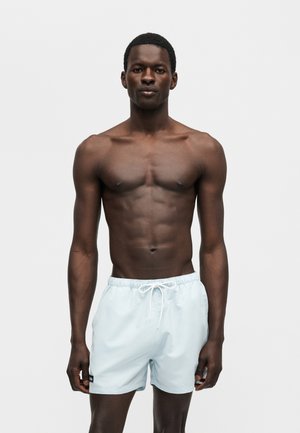 Αθλητικός άνδρας με σκούρο δέρμα, όρθιος και κοιτάζοντας μπροστά, φοράει ανοιχτό μπλε μαγιό με λευκό κορδόνι, σε απλό ανοιχτό φόντο.