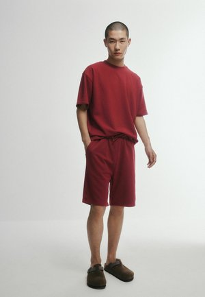 Nuori mies seisoo yksiväristä taustaa vasten, yllään yhteensopiva punainen ylisuurikokoinen T-paita ja shortsit sekä ruskeat slip-on-kengät.