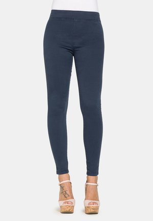 Gambe che indossano pantaloni aderenti blu scuro e sandali rosa chiaro con tacco alto e plateau in sughero, mostrando tatuaggi alla caviglia.