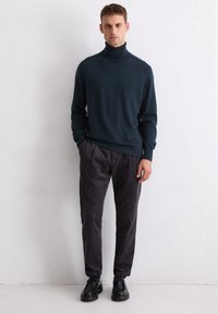 Uomo che indossa un maglione a collo alto di un verde scuro, pantaloni in velluto a coste grigio scuro e scarpe di pelle nere, in piedi contro un muro bianco testurizzato.