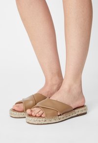 Pieds portant des sandales à enfiler à lanières croisées en cuir beige avec des semelles en jute tressé sur un fond blanc.
