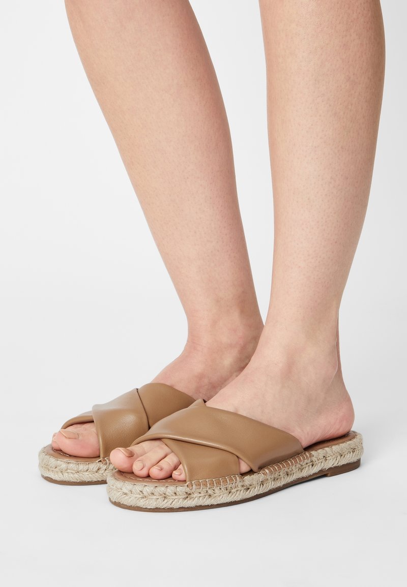 Pieds portant des sandales à enfiler à lanières croisées en cuir beige avec des semelles en jute tressé sur un fond blanc.