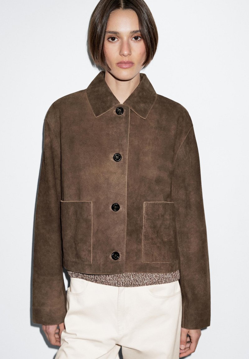 Hnědá semišová bunda s límcem, černými knoflíky, dvěma předními kapsami a krátkým střihem. Materiál má měkkou strukturu.