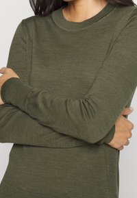 Olivengroene trui met lange mouwen, ronde hals en geribbelde manchetten. De stof is textuurrijk, wat zorgt voor een aansluitende, getailleerde uitstraling.