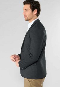 Blazer pour homme gris foncé, design structuré, coupe ajustée, fermeture à un bouton, deux poches avant, tissu lisse. Associé à une chemise blanche.