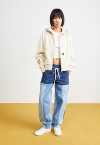 Beige zip-up hoodie, white cropped t-shirt, mixed denim pants with light and dark blue tones, white sneakers, standing on a yellow floor.