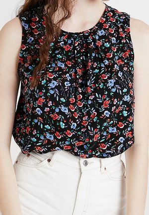 Blouse noire sans manches avec motif floral rouge, bleu et vert, portée rentrée dans un jean blanc taille haute.