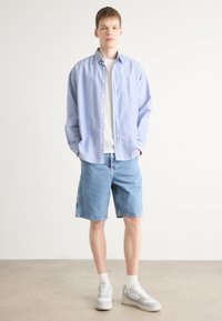 Camisa de botones a rayas azules y blancas sobre una camiseta blanca, combinada con pantalones cortos de mezclilla azul claro y zapatillas grises. Ajuste simple y relajado.