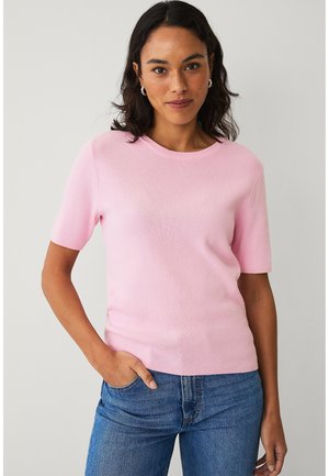 Kvinde med mørkt hår iført en lyserød kortærmet top og blå jeans, stående med den ene hånd i lommen mod en ensfarvet baggrund.