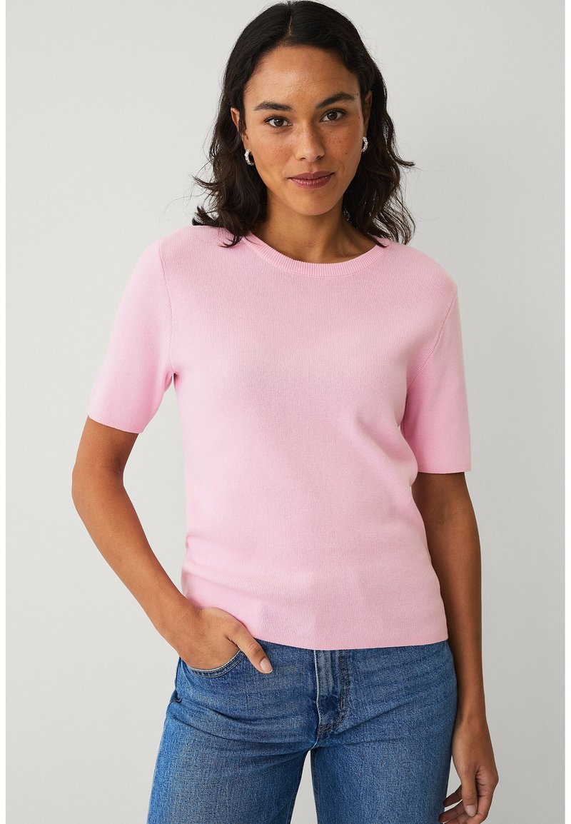 Žena s tmavými vlasy, oblečená v růžovém tričku s krátkými rukávy a modrých džínách, stojící s jednou rukou v kapse na jednobarevném pozadí.