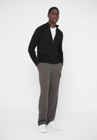 Čierny sveter na zips s textúrovanou látkou, kombinovaný so sivými širokými nohavicami a bielymi teniskami. Jednoduchý dizajn, hladká textúra a neutrálne farby.