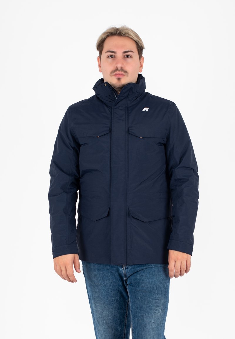Giacca isolante blu navy con colletto alto, chiusura frontale con zip e due tasche sul petto. Presenta un logo sul lato sinistro superiore.