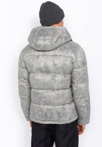Grå quiltet pufferjakke med hætte, der har et tekstureret, akvarel-lignende mønster og elastiske manchetter. Båret med en mørk strikket hue.