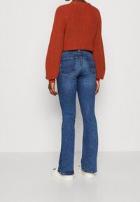 Rostfärgad stickad tröja med ballongärmar, bärs över blå utsvängda jeans. Jeansen har standardfickor och är något urblekta.