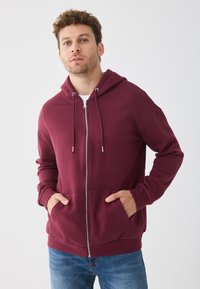 Sweat à capuche bordeaux zippé avec cordon de serrage, poches avant et poignets côtelés. Fabriqué à partir d'un matériau doux et texturé.