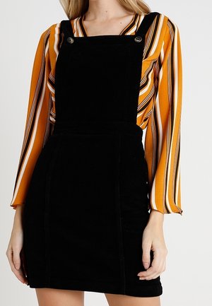 Kobieta ubrana w czarną sukienkę na szelkach z sztruksu nałożoną na bluzkę w pionowe paski w kolorze musztardowym, białym i czarnym z długim rękawem.