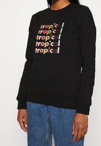Persona que lleva un sudadero negro con la palabra "tropical" impresa cinco veces en letras coloridas, combinado con jeans azules.