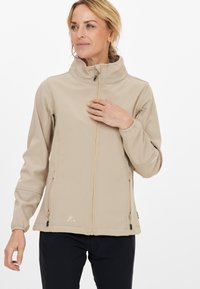 Beige mjukskalsjacka med hög krage, dragkedja fram och två sidfickor. Har elastiska ärmslut och en liten logotyp på bröstet.