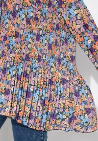 Květinové plisované šaty s kombinací fialových, modrých, oranžových a růžových květin na tmavém pozadí. Mají dlouhé rukávy a rozšířený lem.