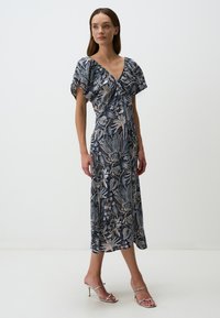 Floral maxi-jurk in marineblauw met korte fluttermouwen, een v-hals en beige accenten. Soepele stof met een vloeiende silhoeët. Zilveren sandalen met hak.