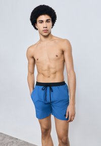 Μπλε μαγιό σορτς με μαύρο ρυθμιζόμενο κορδόνι στη μέση, πλαϊνές τσέπες και μικρή λεπτομέρεια με λογότυπο. Ομαλή υφή και μήκος πάνω από το γόνατο.