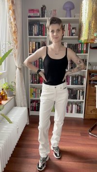 Balti tiesūs džinsai su priekiniais kišenėmis, derinami su juodu marškinėlių topu. Būdingos drąsios raštai ant sportbačių ir knygų lentyna fone.