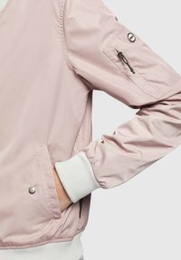 Veste bomber rose clair avec poignets et ourlet côtelés blancs, poche zippée sur la manche, main placée dans la poche latérale.