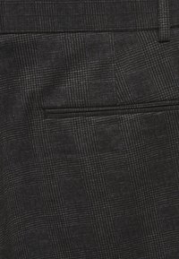 Tissu à carreaux noirs et gris avec une texture de quadrillage subtile, doté d'une poche arrière unique et d'un design épuré et sur mesure.