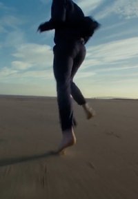 Une personne court pieds nus sur une plage de sable, portant un pantalon sombre et une veste. Le ciel est dégagé avec des nuages légers.